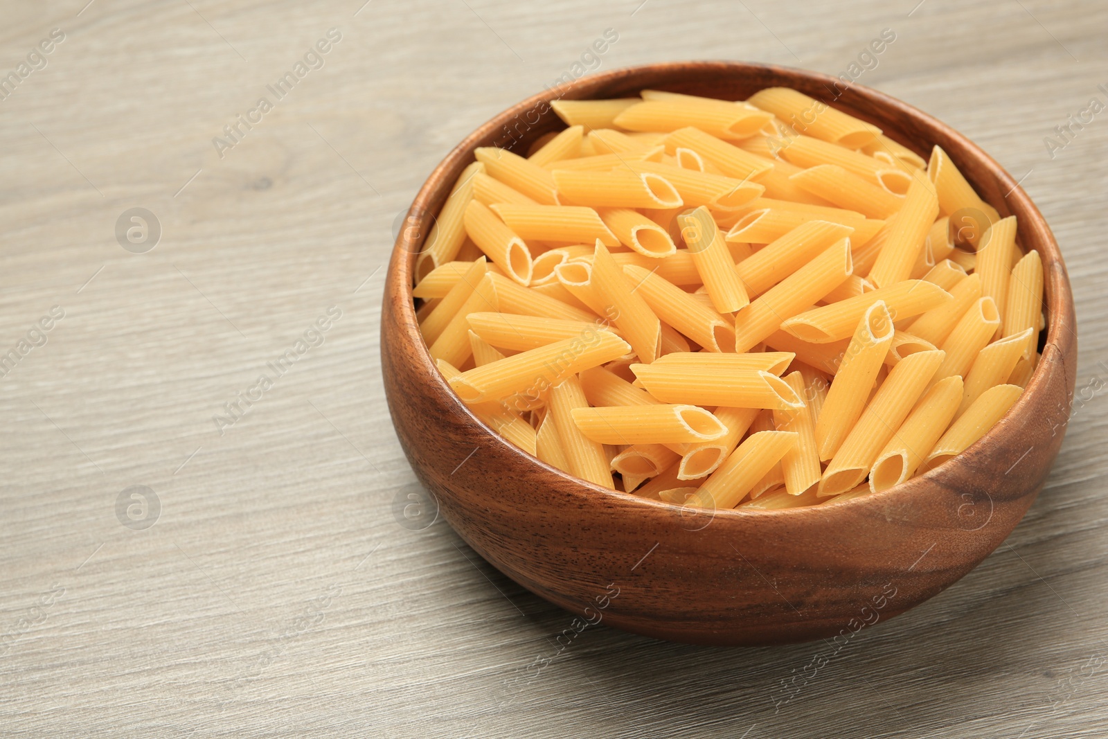 Photo of Raw penne pasta in bowl on light grey wooden table, closeup. Space for text