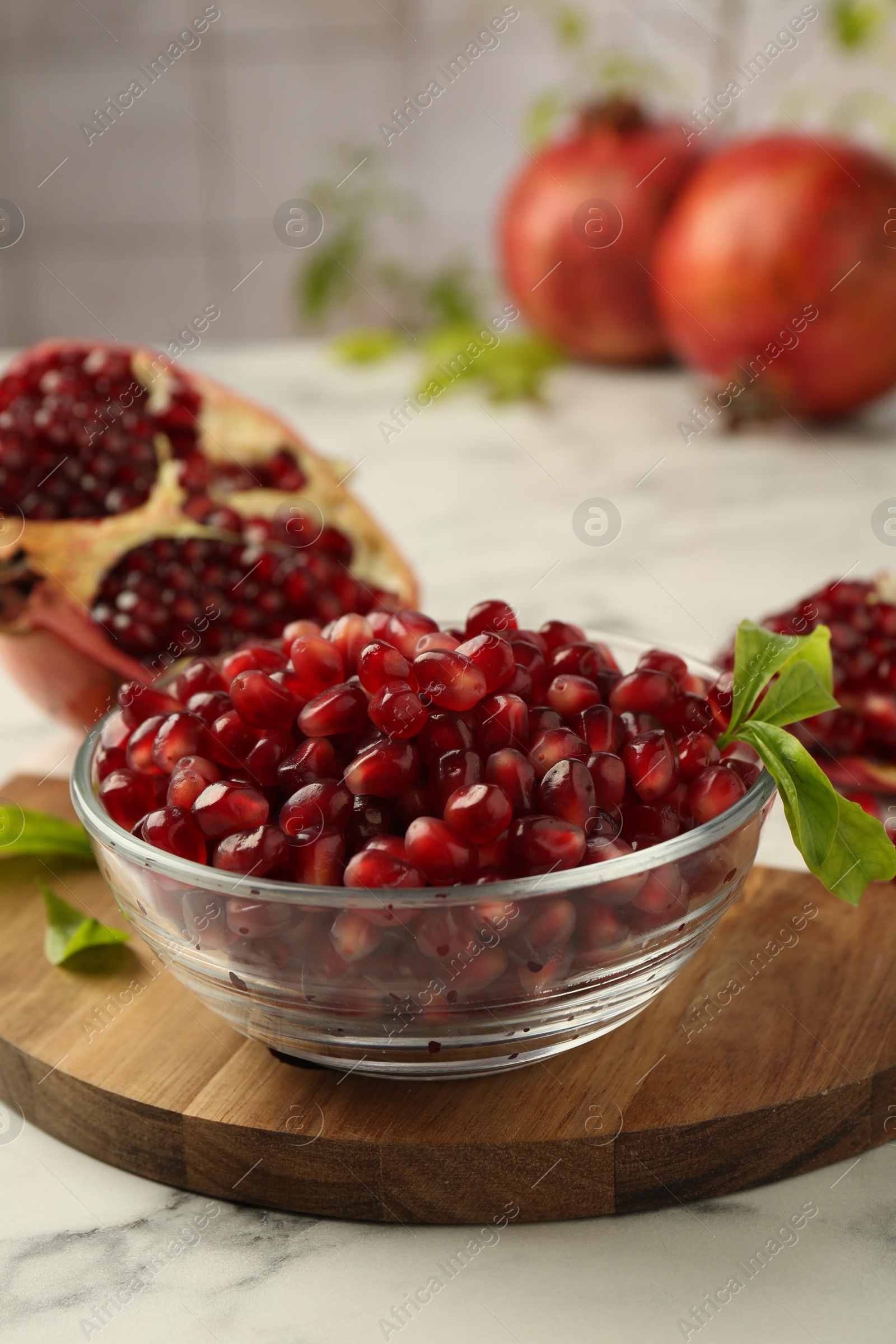 Photo of Ripe juicy pomegranate grains in bowl and green leaves on white marble table