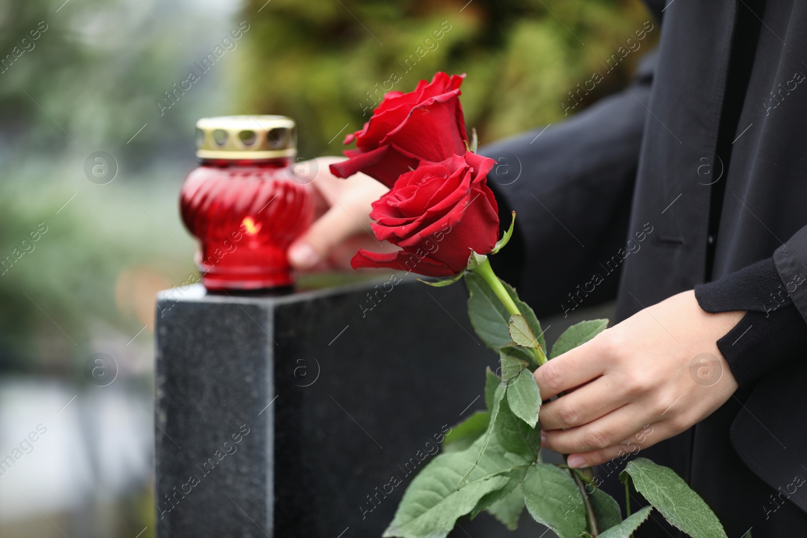 Photo of Woman holding red roses near black granite tombstone with candle outdoors, closeup. Funeral ceremony