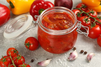 Photo of Glass jar of delicious canned lecho and fresh ingredients on light grey table, closeup