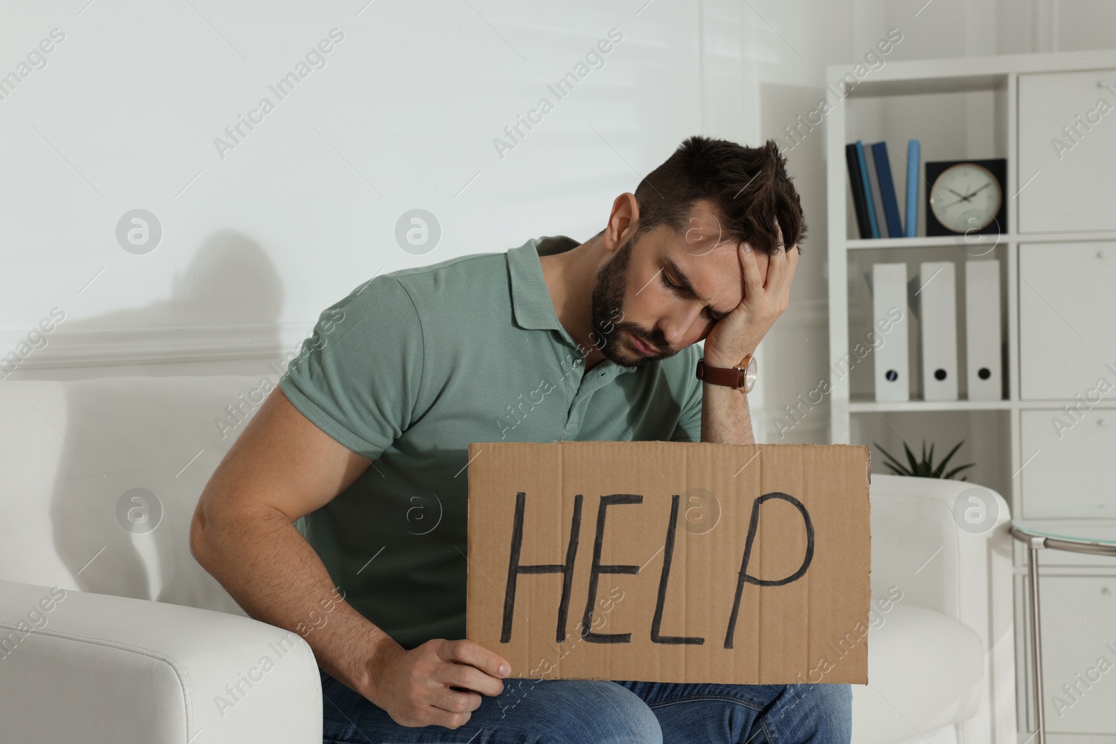 Photo of Unhappy man with HELP sign on sofa indoors