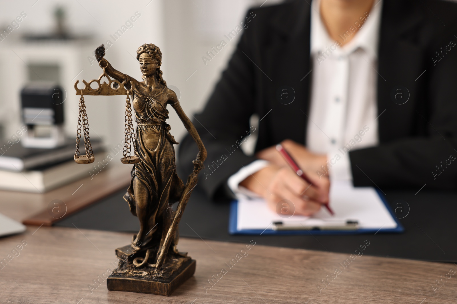 Photo of Notary with clipboard writing notes at workplace in office, focus on statue of Lady Justice