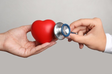 Doctor holding stethoscope near toy heart in patient's hand against grey background, closeup. Pulse checking