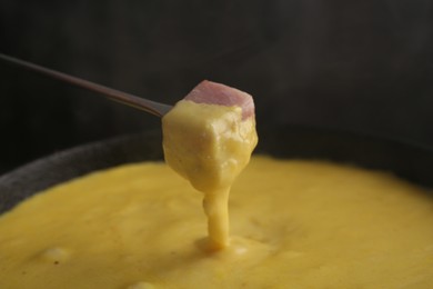 Photo of Dipping piece of ham into fondue pot with tasty melted cheese against dark gray background, closeup