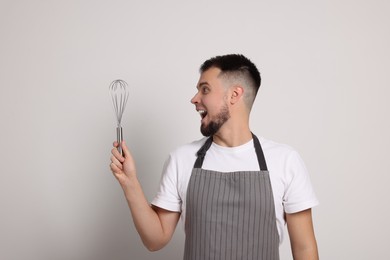 Photo of Surprised professional confectioner in apron holding whisk on light grey background
