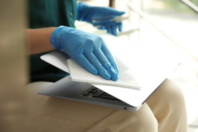 Photo of Woman in latex gloves cleaning laptop with wet wipe indoors, closeup