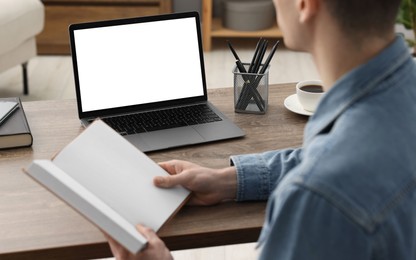 Photo of E-learning. Young man with book during online lesson at table indoors, closeup