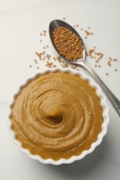 Photo of Tasty mustard sauce in bowl and spoon with dry seeds on white table, flat lay