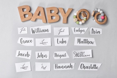 Photo of Paper sheets with written different baby names, wooden letters and toys on grey background, flat lay