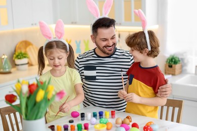 Photo of Easter celebration. Father and his cute children with bunny ears painting eggs at white marble table in kitchen