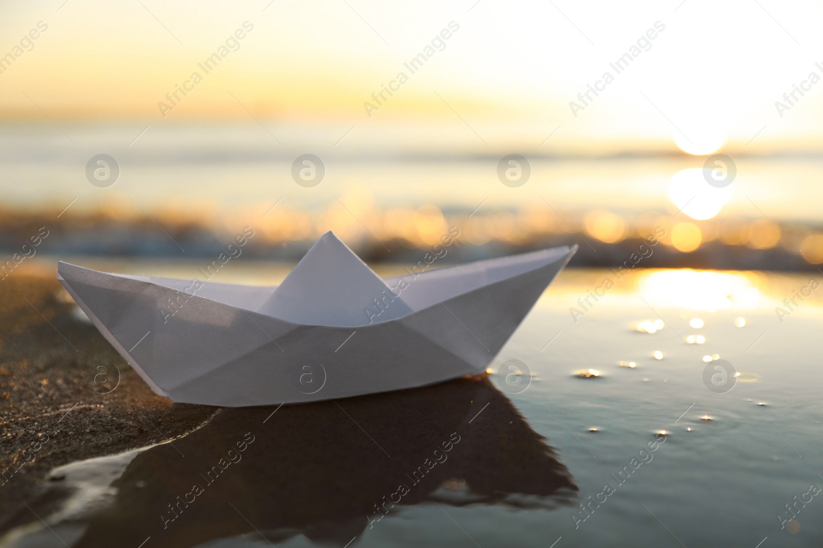 Photo of White paper boat on wet sand near sea at sunset, space for text