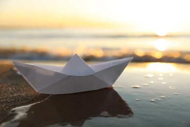 Photo of White paper boat on wet sand near sea at sunset, space for text