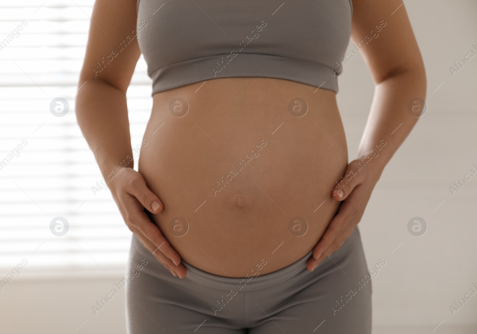 Photo of Pregnant young woman touching belly near window indoors, closeup