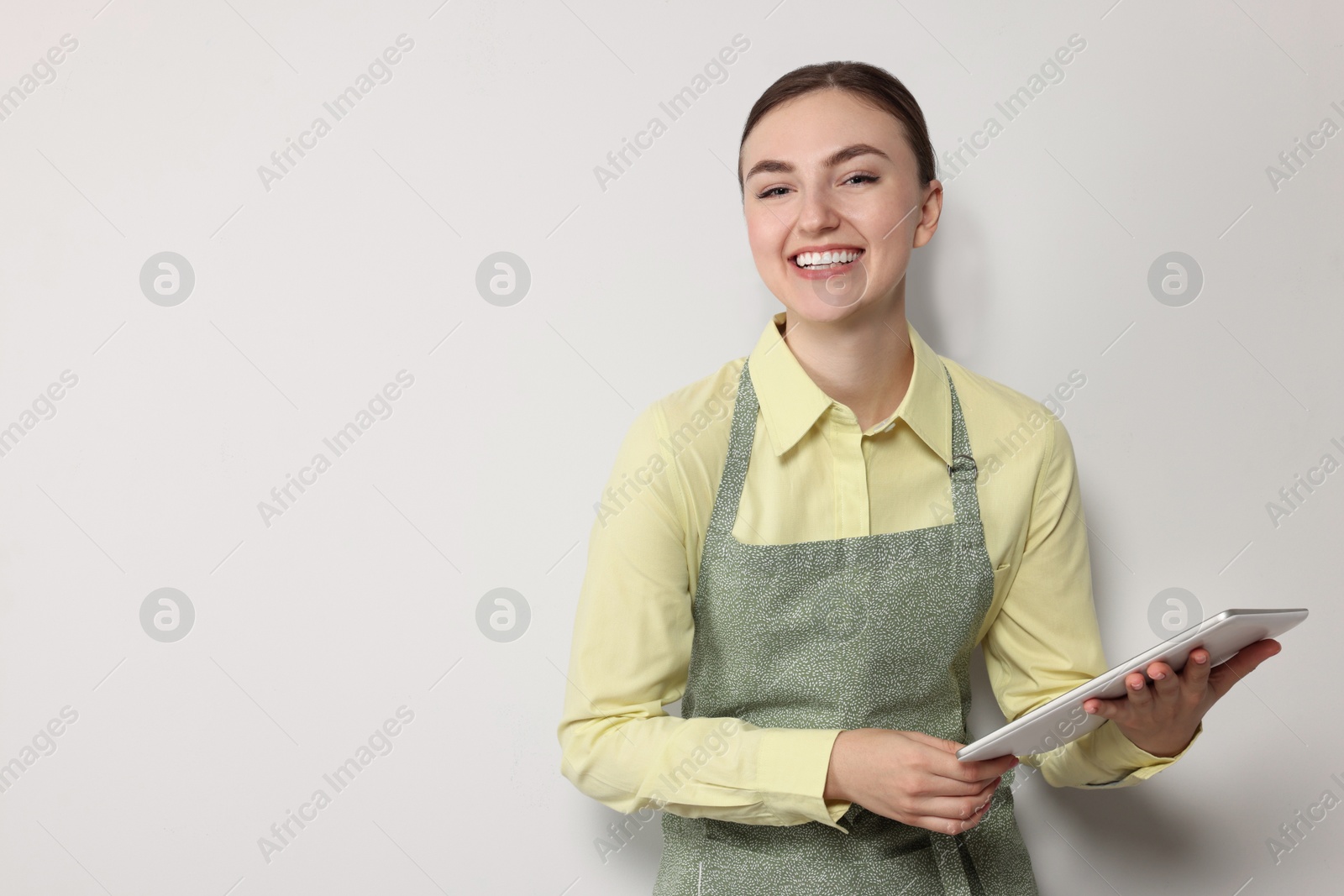 Photo of Beautiful young woman in clean apron with tablet on light grey background. Space for text