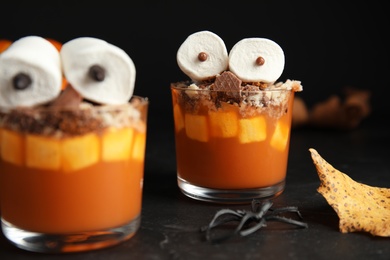 Photo of Glasses with delicious dessert decorated as monsters on black table, closeup. Halloween treat