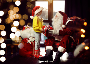 Photo of Santa Claus giving Christmas gift to little girl near window indoors