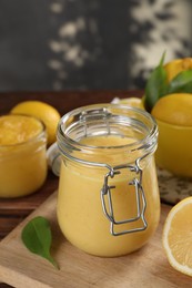 Photo of Delicious lemon curd in glass jar, fresh citrus fruits and green leaves on table