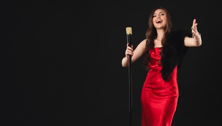 Beautiful young woman in stylish dress with microphone singing on black background