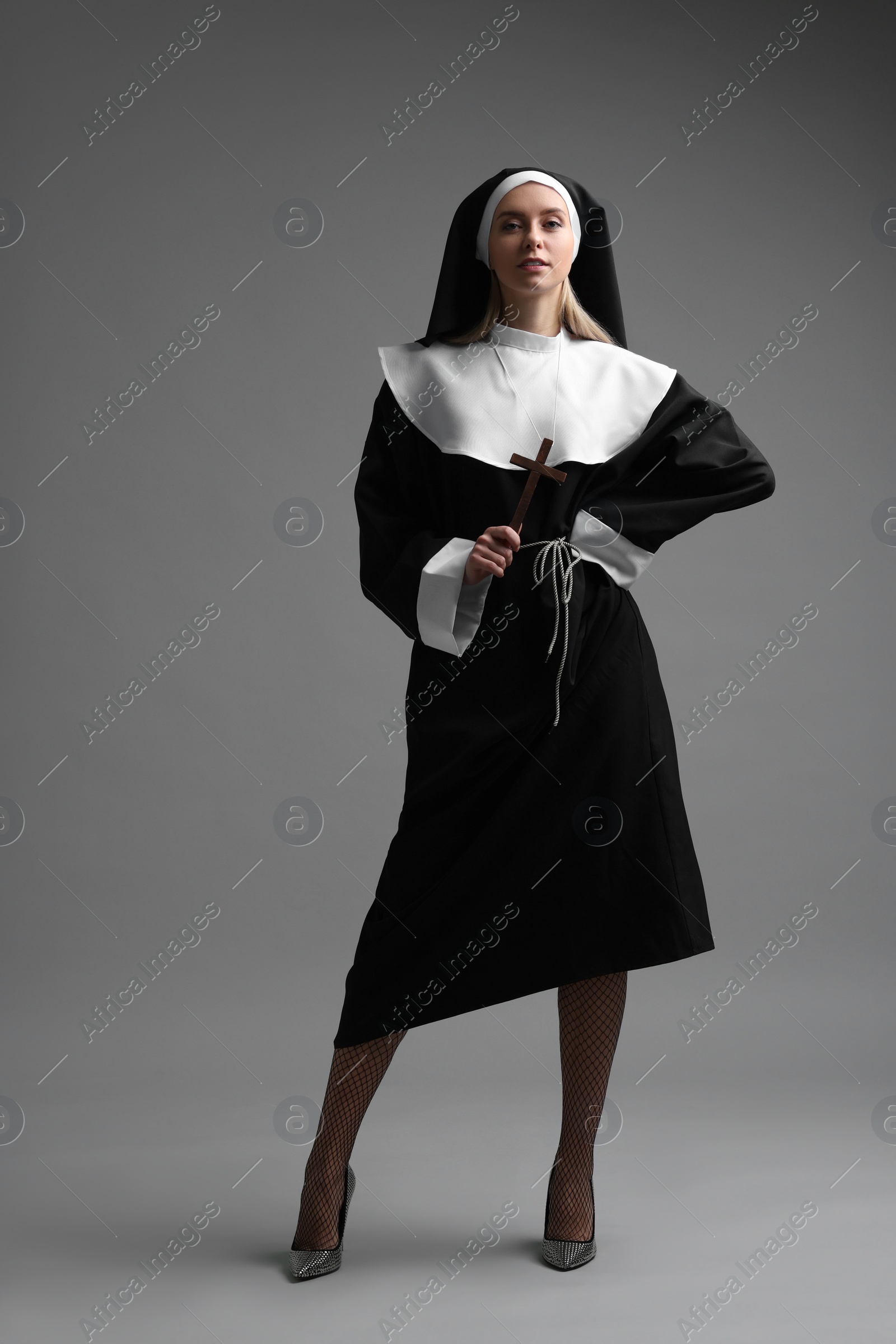 Photo of Woman in nun habit and mesh tights holding wooden cross on grey background. Sexy costume