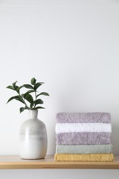 Photo of Stacked terry towels and green branches in vase on wooden shelf near white wall