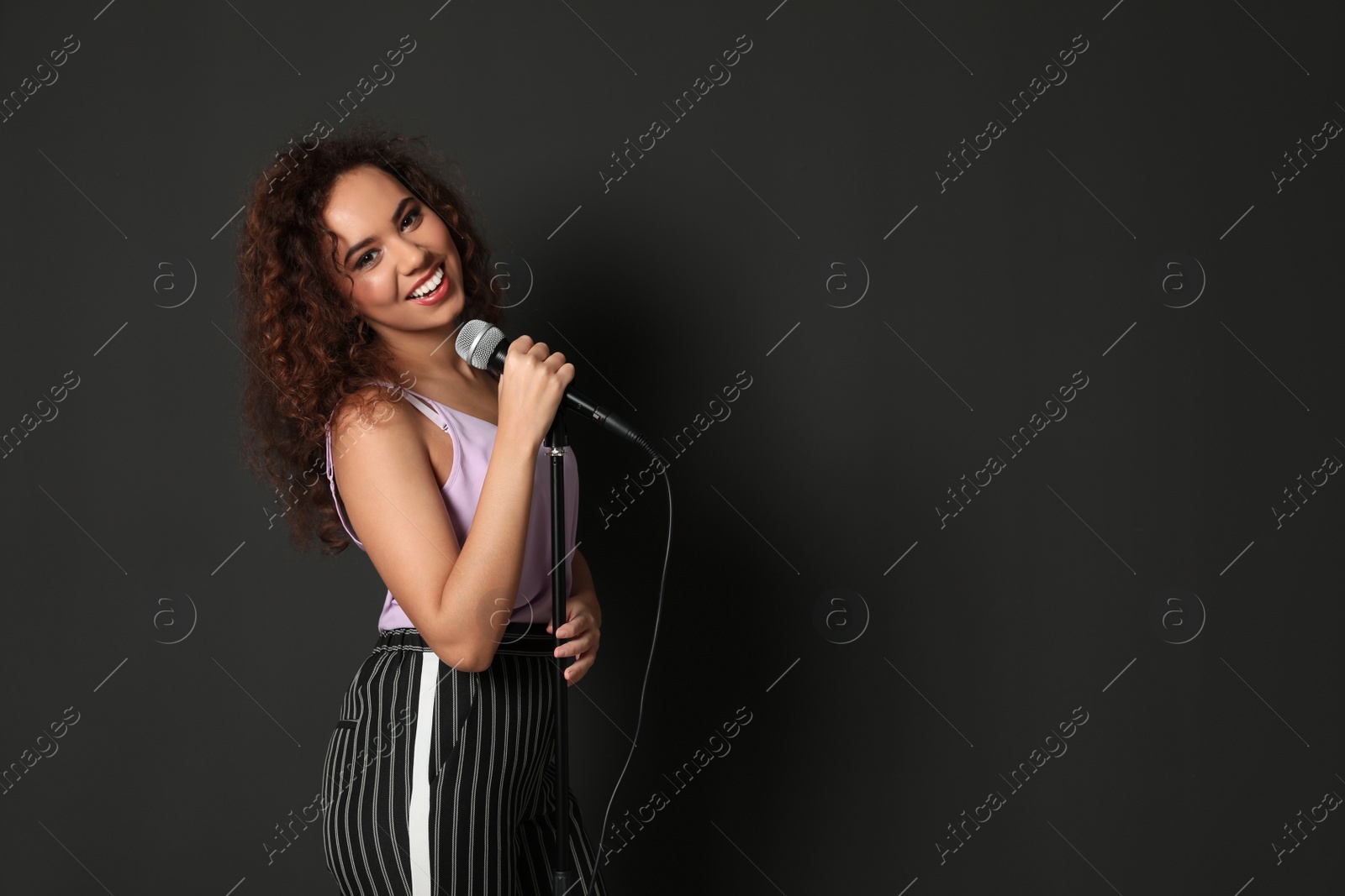 Photo of Curly African-American woman in stylish clothes posing with microphone on black background. Space for text