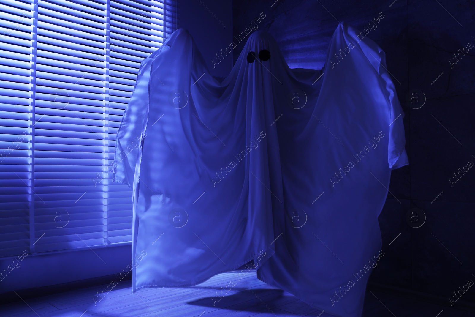 Photo of Creepy ghost. Woman covered with sheet near window in blue light