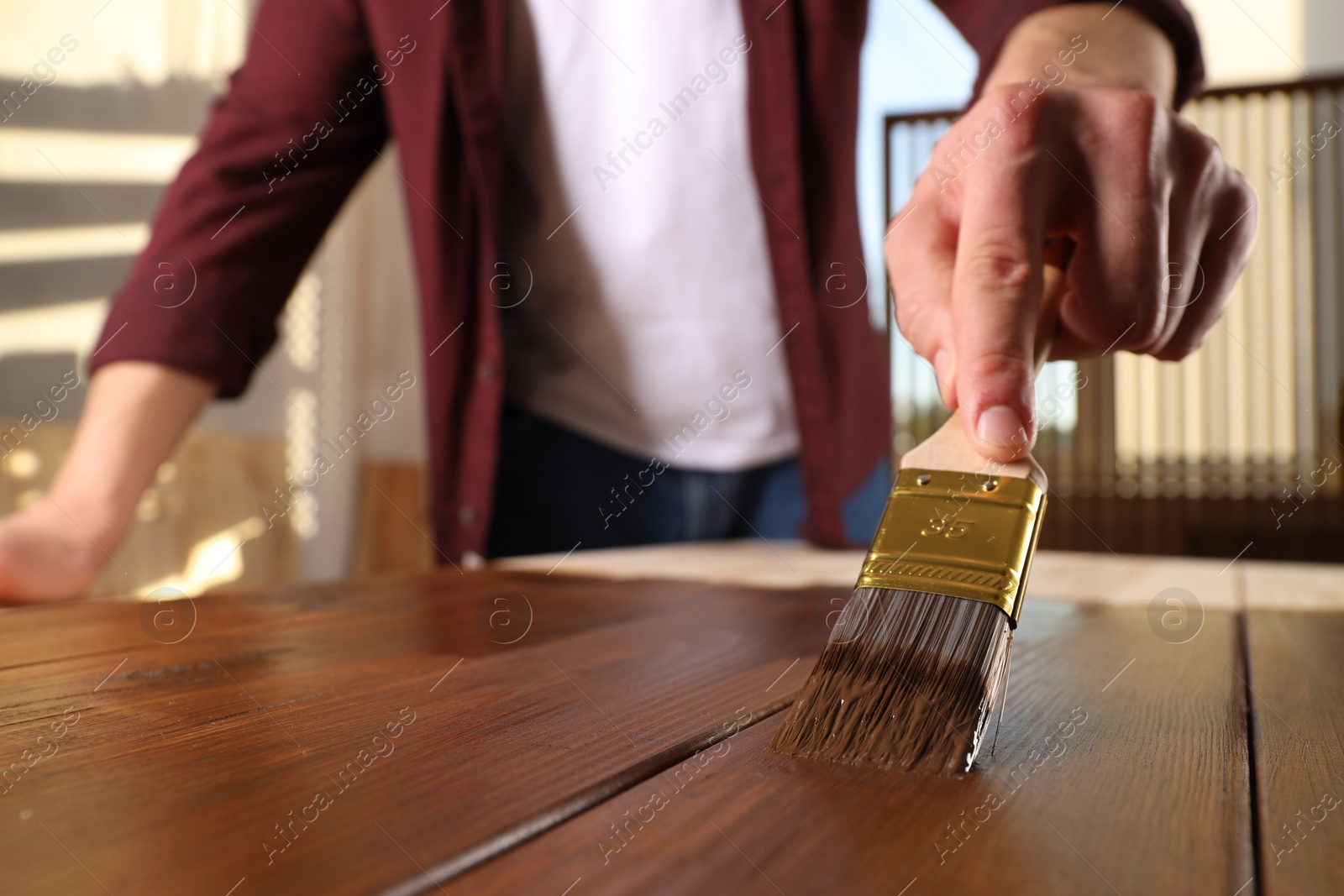 Photo of Man with brush applying wood stain onto wooden surface indoors, closeup