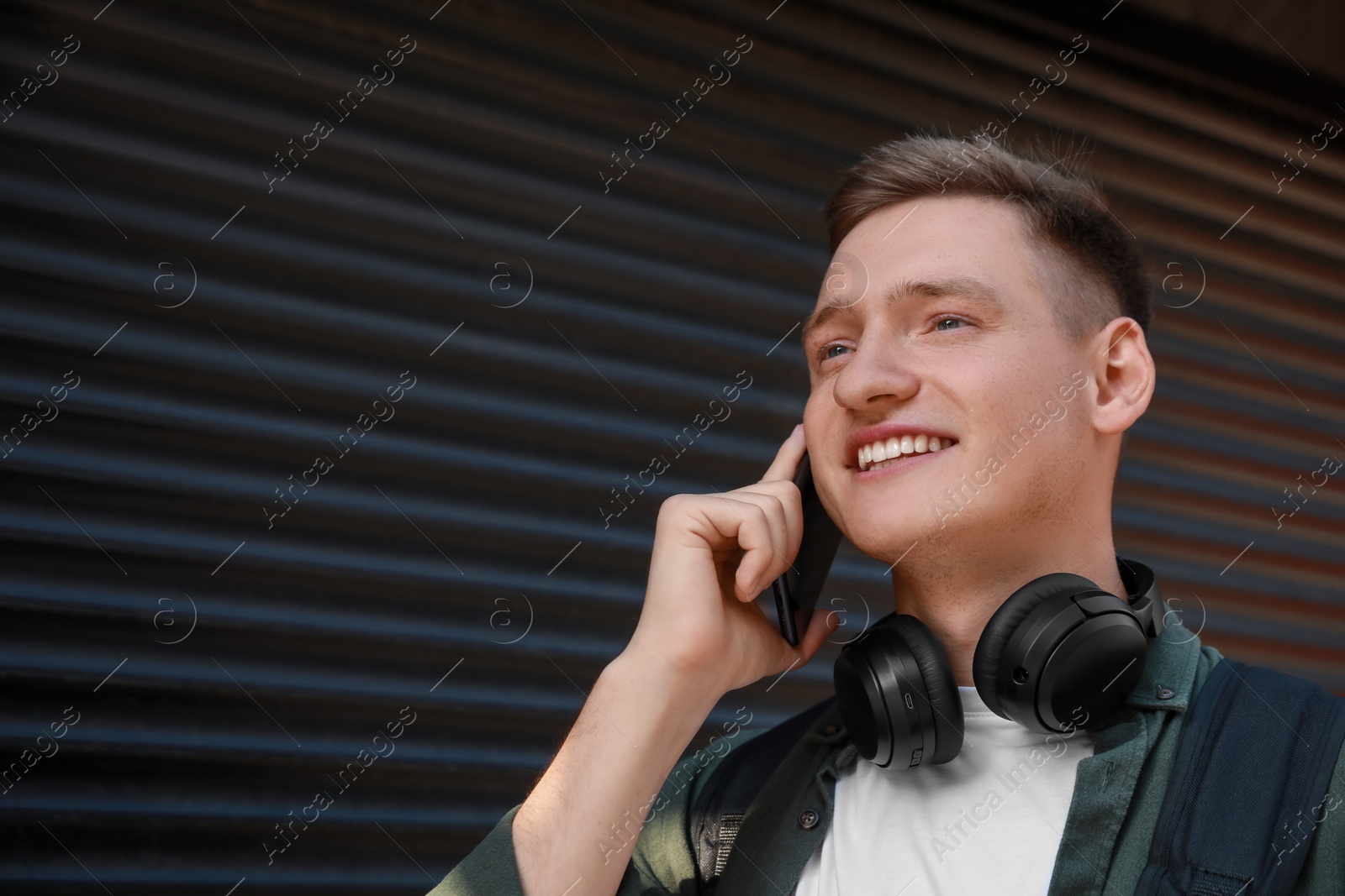 Photo of Smiling man with headphones talking on smartphone near shutters outdoors. Space for text
