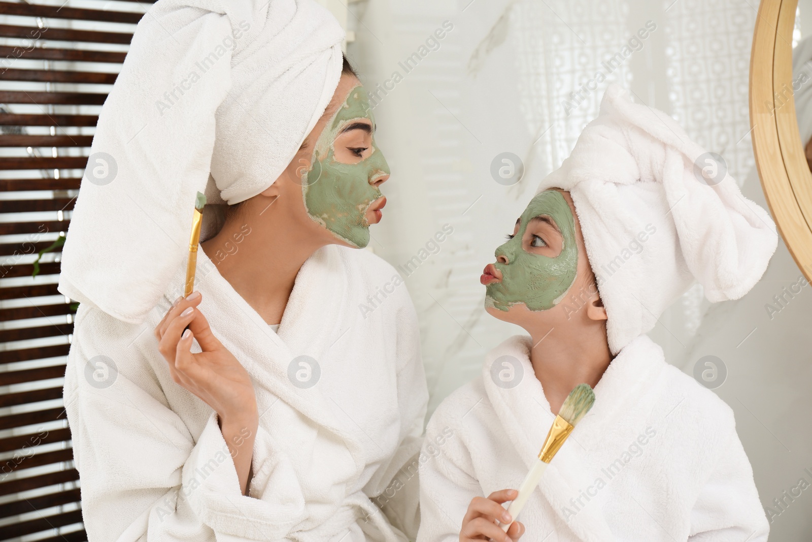 Photo of Young mother and her daughter with facial masks having fun in bathroom