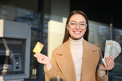 Happy young woman with credit card and money near cash machine outdoors