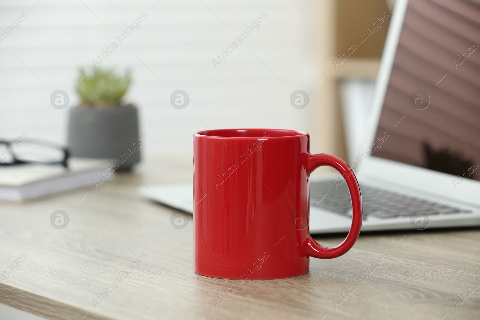 Photo of Red ceramic mug and laptop on wooden table indoors