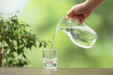 Photo of Woman pouring fresh water from jug into glass at wooden table against blurred green background, closeup. Space for text