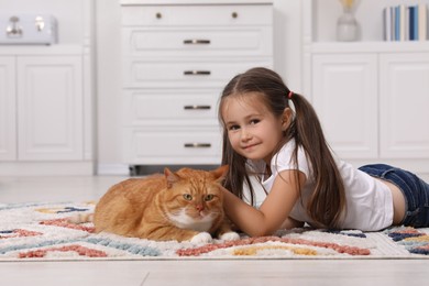 Smiling little girl petting cute ginger cat on carpet at home