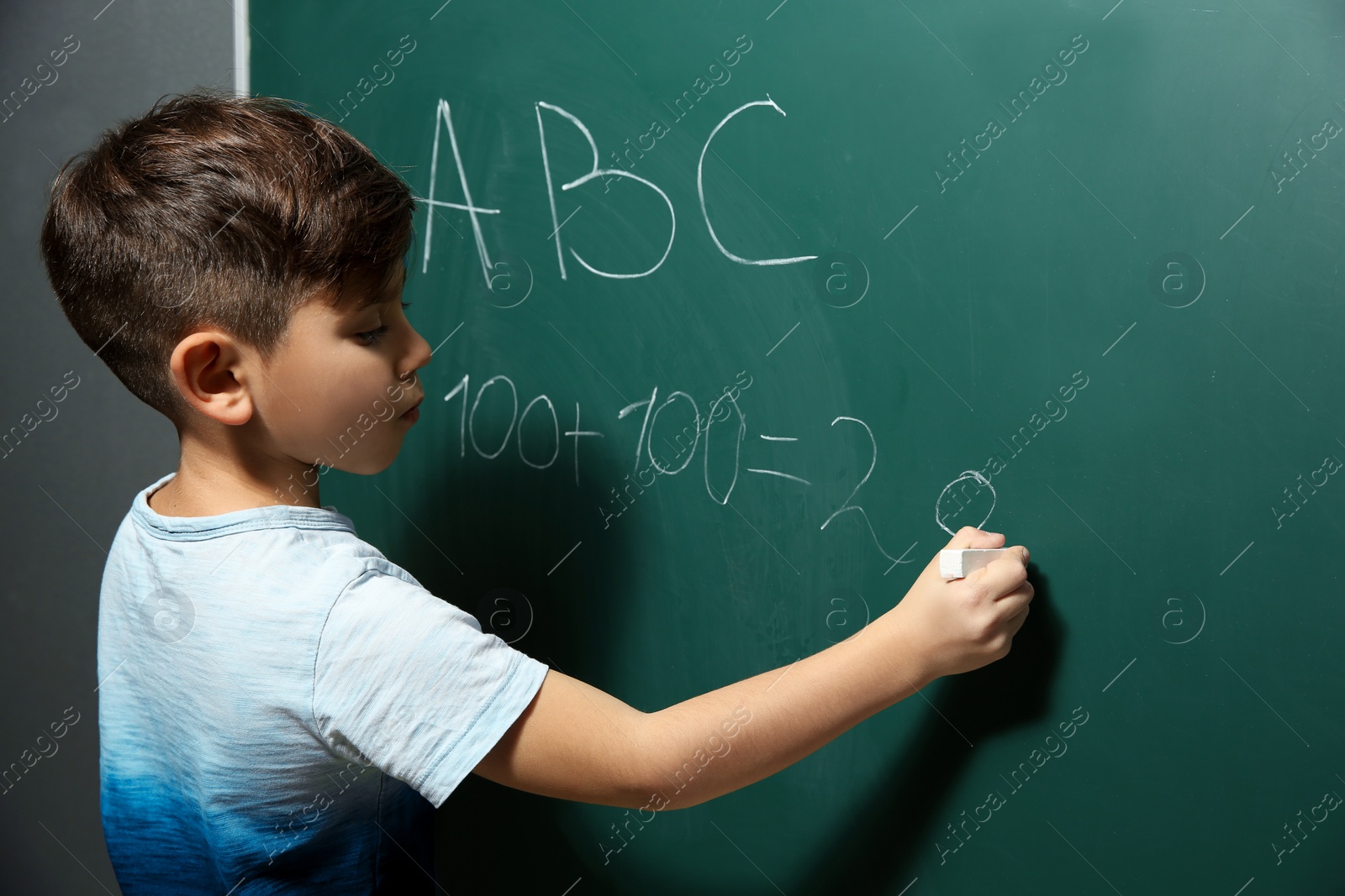 Photo of Little child writing letters and doing math on green blackboard