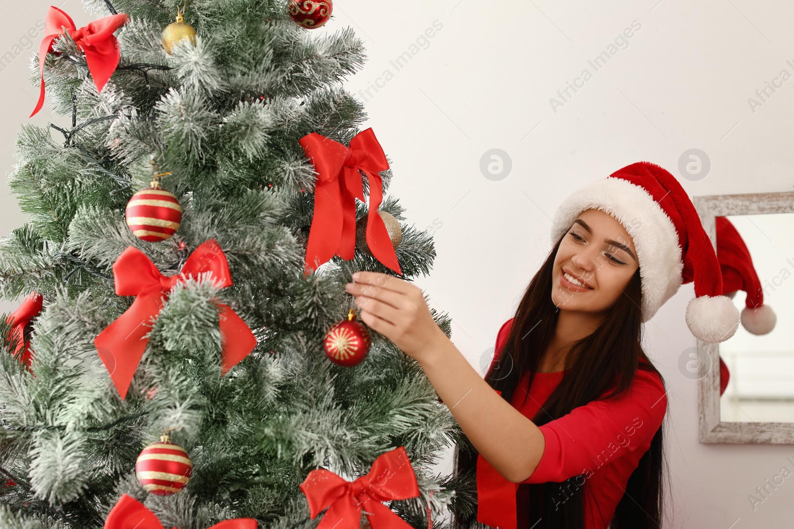 Photo of Beautiful young woman in Santa hat decorating Christmas tree at home