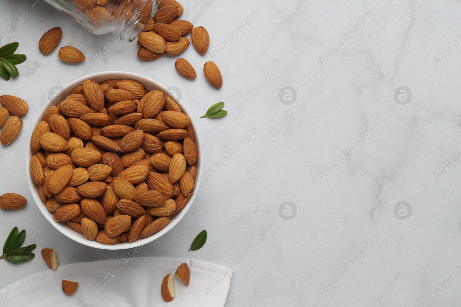 Photo of Bowl with delicious almonds and fresh leaves on white marble table, flat lay. Space for text