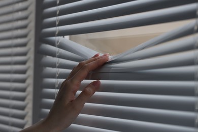 Woman separating slats of white blinds indoors, closeup