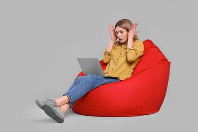 Emotional woman with laptop sitting on beanbag chair against light grey background