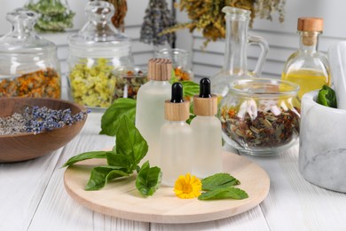 Bottles of essential oils, leaves, flower and herbs on white wooden table