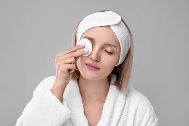 Young woman cleaning her face with cotton pad on light grey background
