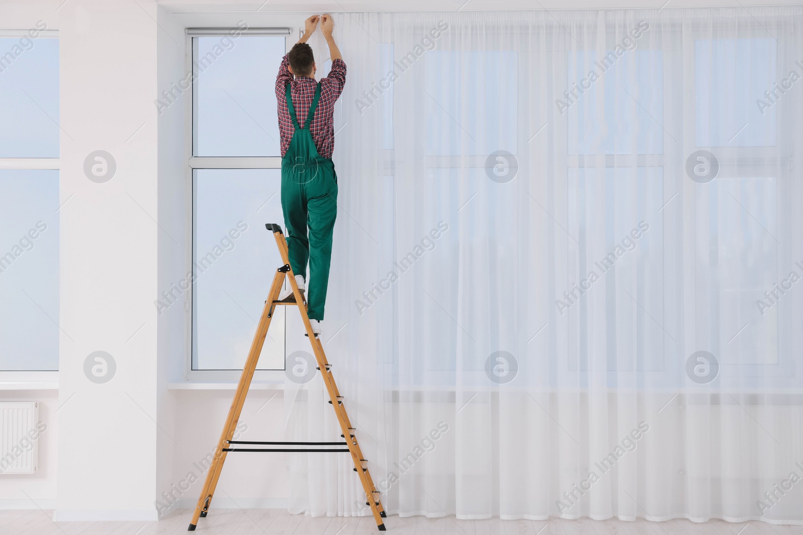 Photo of Worker in uniform hanging window curtain indoors, back view