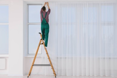 Worker in uniform hanging window curtain indoors, back view