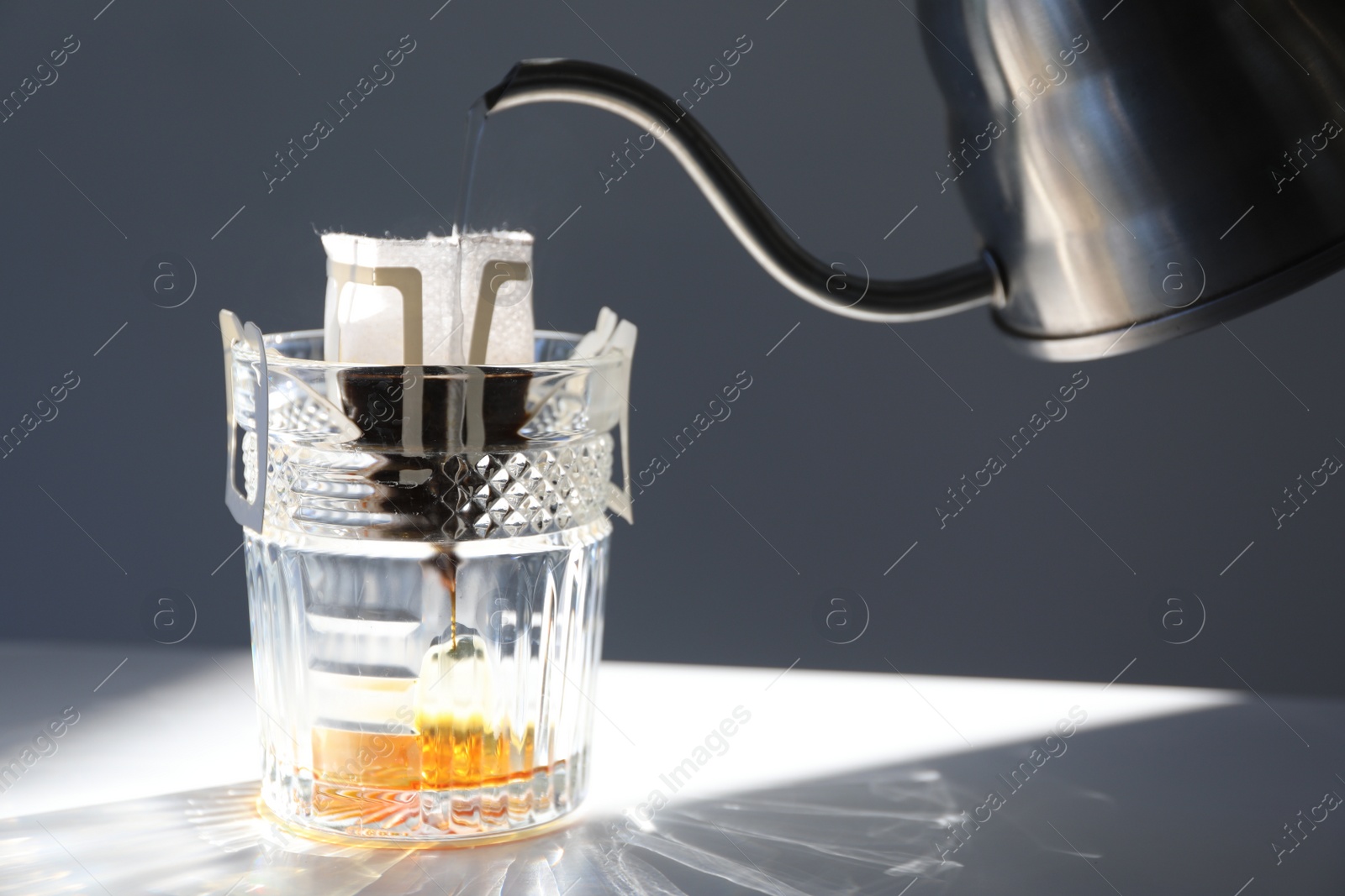 Photo of Pouring hot water into glass with drip coffee bag from kettle on light grey table, closeup