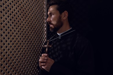Photo of Catholic priest in cassock holding cross in confessional booth, space for text