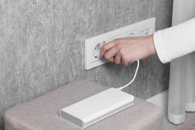 Photo of Woman plugging power bank into socket on grey wall indoors, closeup