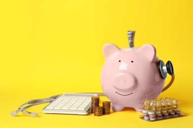 Photo of Piggy bank with money, stethoscope, calculator and pills on yellow background, space for text. Medical insurance