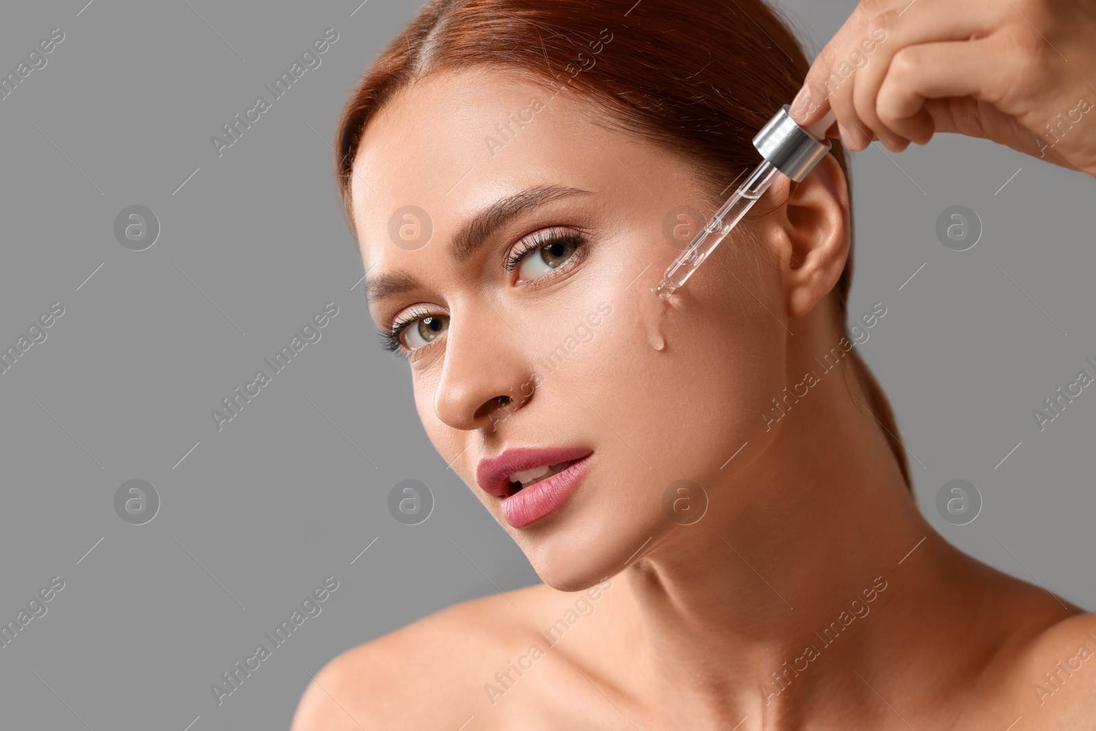 Photo of Beautiful young woman applying cosmetic serum onto her face on grey background, space for text