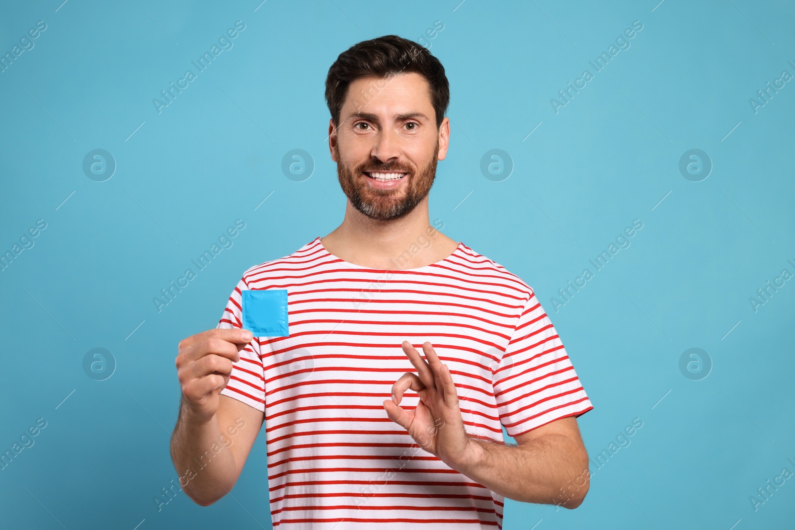 Photo of Happy man with condom showing ok gesture on light blue background. Safe sex