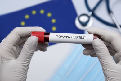 Photo of Doctor holding sample tube with label Coronavirus Test above medical items and European Union flag, closeup