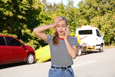 Photo of Woman talking on phone near broken car and tow truck outdoors